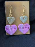 gold heart dangling above pink heart that says cunt