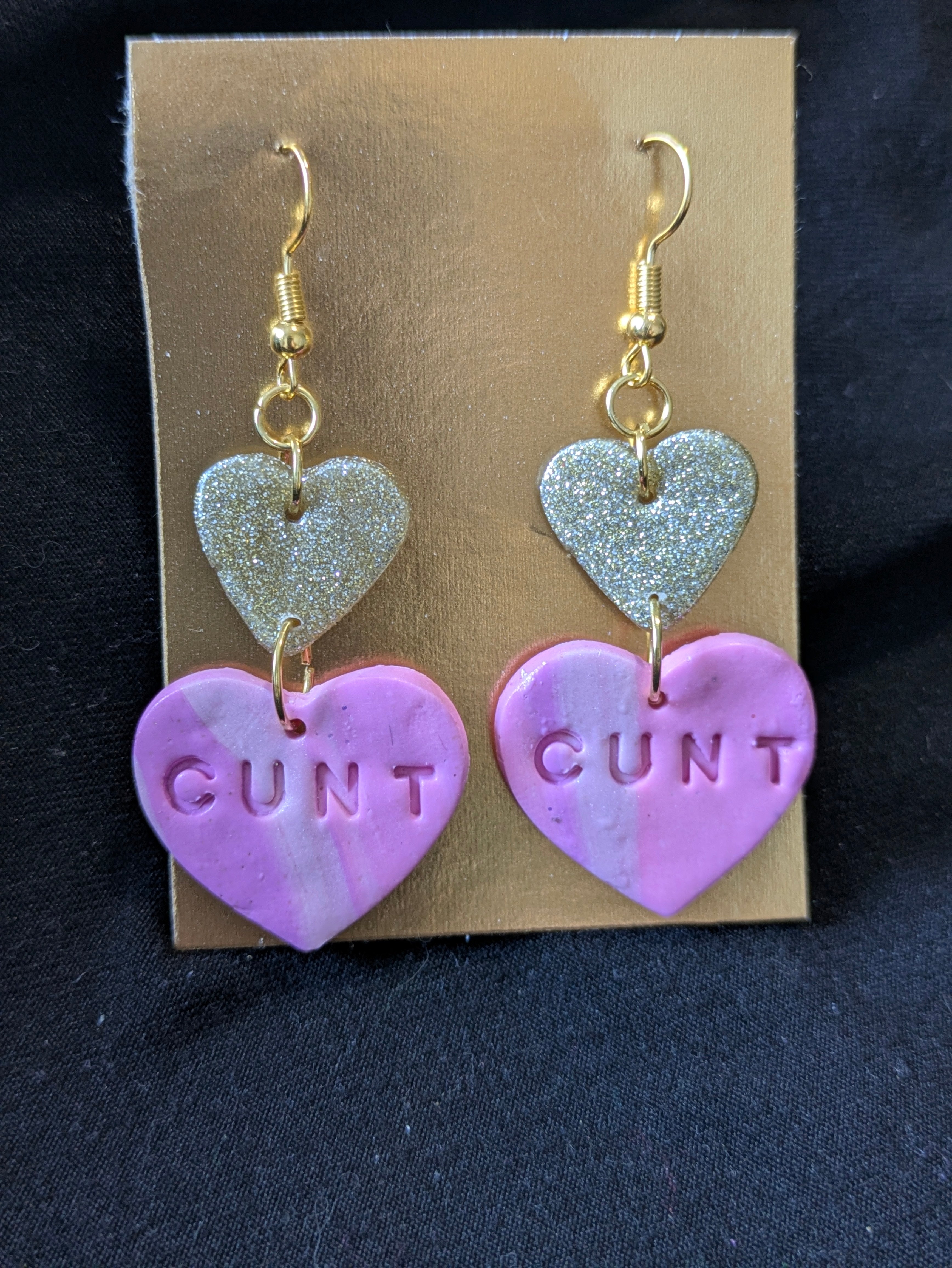gold heart dangling above pink heart that says cunt